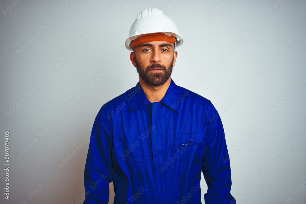 Handsome indian worker man wearing uniform and helmet over isolated white background Relaxed with serious expression on face. Simple and natural looking at the camera. - obrazy, fototapety, plakaty 