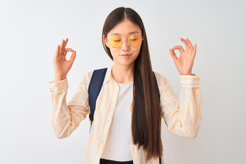 Sticker - Young chinese student woman wearing glasses and backpack over isolated white background relax and smiling with eyes closed doing meditation gesture with fingers. Yoga concept.