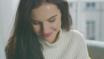 Wall Mural - Portrait of Brunette Smiling Charmingly while Sitting on Sofa, Brushes Her Lush Hair Away with Flirtatious Gesture. Girl Wearing Oversized White Knitted Sweater Relaxes on Couch in Her Cozy Apartment