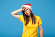 © ViDi Studio - Exhausted young brunette woman Santa girl in yellow t-shirt Christmas hat posing isolated on blue wall background. New Year 2020 celebration holiday concept. Mock up copy space. Putting hand on head.