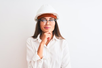 Poster - Young beautiful architect woman wearing helmet and glasses over isolated white background looking confident at the camera with smile with crossed arms and hand raised on chin. Thinking positive.