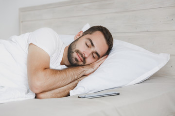 Wall Mural - Calm young handsome bearded man lying in bed with white sheet pillow blanket in bedroom at home. Smiling beauty male spending time in room. Rest relax good mood lifestyle concept. Mock up copy space.