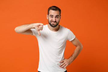 Displeased confused young man in casual white t-shirt posing isolated on orange wall background studio portrait. People sincere emotions lifestyle concept. Mock up copy space. Showing thumb down.