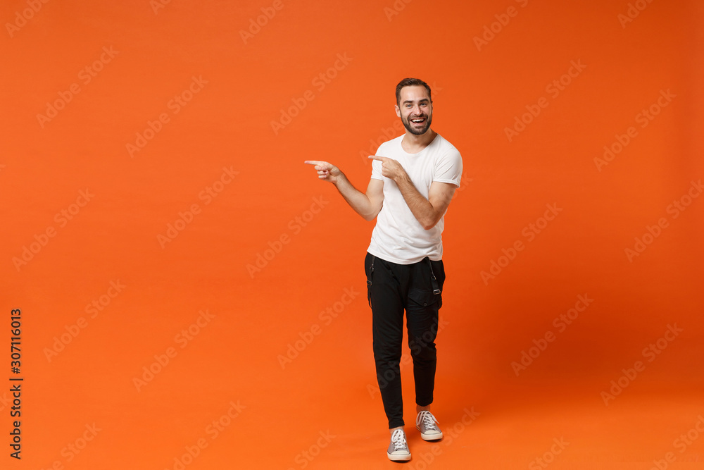 Smiling young man in casual white t-shirt posing isolated on bright orange wall background studio portrait. People sincere emotions lifestyle concept. Mock up copy space. Pointing index fingers aside. - obrazy, fototapety, plakaty 
