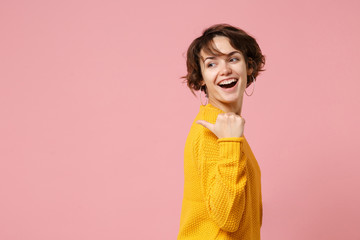 Wall Mural - Side view of laughing young brunette woman girl in yellow sweater posing isolated on pastel pink wall background studio portrait. People lifestyle concept. Mock up copy space. Pointing thumb aside.