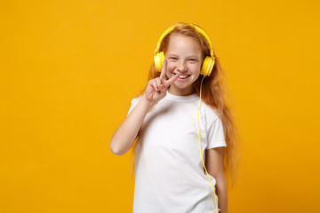 Wall Mural - Little ginger kid girl 12-13 years old in white t-shirt isolated on yellow background studio portrait. Childhood lifestyle concept. Mock up copy space. Listen music in headphones showing victory sign.