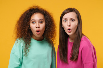 Two shocked women friends european african american girls in pink green clothes posing isolated on yellow background studio portrait. People lifestyle concept. Mock up copy space. Keeping mouth open.