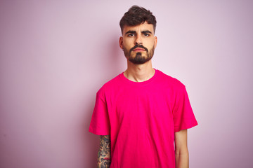 Sticker - Young man with tattoo wearing t-shirt standing over isolated pink background with serious expression on face. Simple and natural looking at the camera.