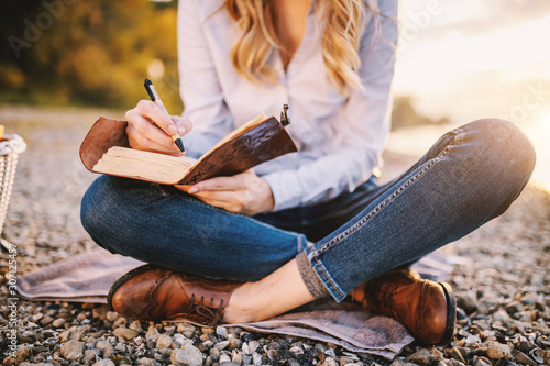Fotografía Cropped picture of focused fashionable beautiful caucasian woman sitting near river and holding notebook