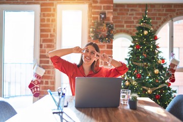 Poster - Beautiful woman sitting at the table working with laptop at home around christmas tree Doing peace symbol with fingers over face, smiling cheerful showing victory