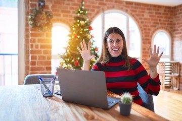 Poster - Beautiful woman sitting at the table working with laptop at home around christmas tree showing and pointing up with fingers number ten while smiling confident and happy.