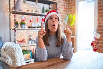 Poster - Young beautiful woman wearing christmas hat sitting at the table at home amazed and surprised looking up and pointing with fingers and raised arms.