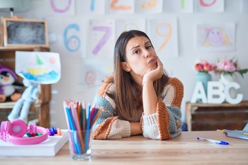 Sticker - Young beautiful teacher woman wearing sweater and glasses sitting on desk at kindergarten thinking looking tired and bored with depression problems with crossed arms.