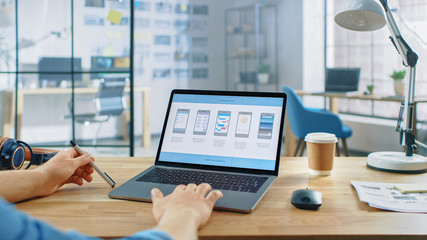 Young Professional Works on a Laptop at His Desk in the Creative Office. Notebook Screen Shows Mobile Phone Application Design, Software UI Development. In the Background Creative Modern Office
