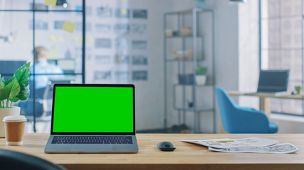 Wall Mural - laptop on the desk in the office shows green mock-up screen. in the background creative office or bu