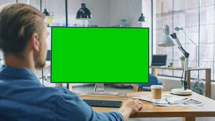 Wall Mural - over the shoulder shot: man uses desktop computer with green mock-up screen while sitting at his des