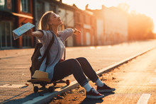 Smiling Girl Sitting On Long Board In The City During Sunset With Arms Outstretched.