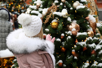 Wall Mural - Girl in a knitted hat and winter coat with fur collar enjoys the snow and magic of Christmas holidays standing on New Year trees background