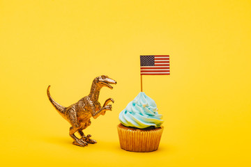 Wall Mural - Toy dinosaur beside cupcake with american flag on yellow background