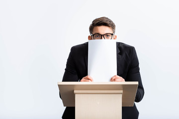 Wall Mural - scared businessman in suit standing at podium tribune and obscuring face with paper during conference isolated on white