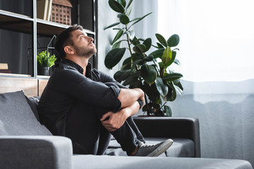 Wall Mural - side view of handsome man with panic attack sitting on sofa and hugging legs in apartment