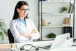 © LIGHTFIELD STUDIOS - female doctor with crossed arms having online consultation with patient on laptop in clinic office