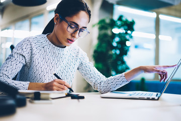 Pensive business woman noting idea for startup planning from web page on laptop computer,professional administrative manager making accountings of income using app on netbook in office interior