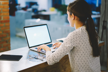Back view of female employee typing on laptop computer with mock up screen downloading multimedia and searching searching information online; woman sitting at working place in office using netbook