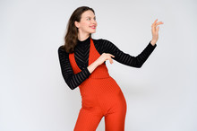 Concept Adult Girl On A White Background. A Photo Of A Pretty Brunette Girl In Red Trousers And A Black Sweater Smiles And Shows Different Emotions In Different Poses Right In Front Of The Camera.