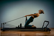 © Georgii - Young girl doing pilates exercises with a reformer bed. Beautiful slim fitness trainer on reformer gray background, low key, art light. Fitness concept