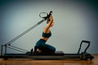© Georgii - Young girl doing pilates exercises with a reformer bed. Beautiful slim fitness trainer on reformer gray background, low key, art light. Fitness concept