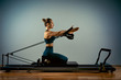 © Georgii - Young girl doing pilates exercises with a reformer bed. Beautiful slim fitness trainer on reformer gray background, low key, art light. Fitness concept