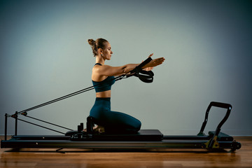 Young girl doing pilates exercises with a reformer bed. Beautiful slim fitness trainer on reformer gray background, low key, art light. Fitness concept