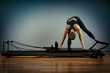 © Georgii - Young girl doing pilates exercises with a reformer bed. Beautiful slim fitness trainer on reformer gray background, low key, art light. Fitness concept
