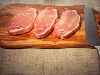 Three fresh uncooked raw port chops on a wooden cutting board. Simple white table cloth.