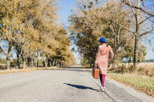 Young Redhead Lady Woman In Pink Vintage Coat And Hat With Suitcase In Retro Style Walking Away Along A Park Road With Golden Yellow Autumnal Trees. Outdoor Autumn Garden Relaxation, Travel Concept