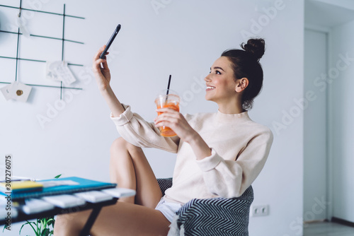 Fotografía Smiling woman taking selfie with smartphone while sitting in armchair at home