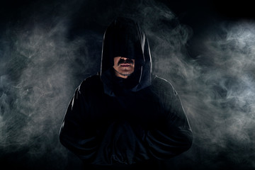 Man dressed in a dark robe looking like a cult leader on a smoky or foggy background.  He looks like a creepy evil villain