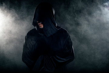 Man dressed in a dark robe looking like a cult leader on a smoky or foggy background.  He looks like a creepy evil villain