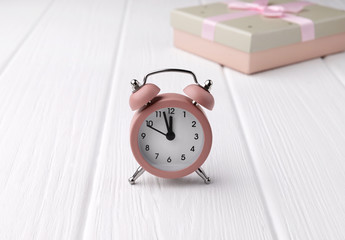 Retro style pink alarm clock amd gift box on white wooden background shows two minutes to twelve o'clock