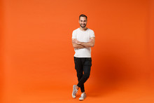 Smiling Attractive Young Man In Casual White T-shirt Posing Isolated On Orange Wall Background Studio Portrait. People Sincere Emotions Lifestyle Concept. Mock Up Copy Space. Holding Hands Crossed.