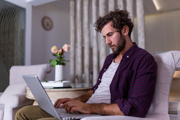 Wall Mural - Man using laptop computer, working at home. Worried about finances. Handsome man with beard working at home on project, he is sitting on sofa looking at his laptop in front of him. Focus on the man