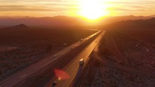 AERIAL, LENS FLARE: Flying Above Trucks And Cars Crossing The Mojave Desert At Sunset. Freight Trucks And Cars Move Along The Asphalt Highway Running Through The California Wilderness On Sunny Evening