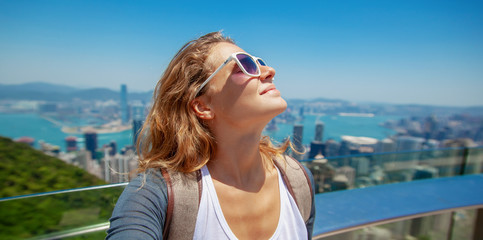 Wall Mural - Beautiful young woman smiles happily and sets her face to the sun on the background of skyscrapers in Hong Kong, traveling to Asia