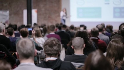 adult people at conference listen to woman speaker providing lecture on scene in big conference hall