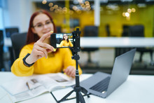 Hand Woman Holding Camera For Recording Video While Sitting At Office. Teenager Student In A Yellow Sweater With Laptop Having Fun Vlogging Live Feeds On Social Media. Technology And Videoblog Concept