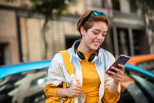 Modern Teen Female In Yellow Trendy Clothes With Wireless Headphones And Yellow Handbag Surfing Mobile Phone While Sitting On Fence Against Colorful Cars And Blurred Modern Buildings In City