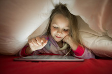 Cute Little Blonde Girl In Pajama Using Tablet And Watching Favorite Cartoons While Lying Under Blanket In Bed At Night