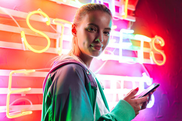 Wall Mural - Image of beautiful woman holding smartphone over neon text sign