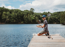 Boy Holding A Fish That He Caught On The End Of A Dock On A Lake.